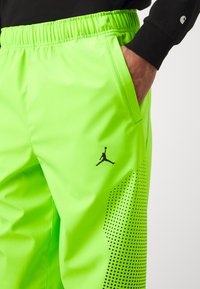 Fluorescenčne zelené športové nohavice s elastickým pásom, bočnými vreckami a čiernym logom Jumpman. Obsahujú bodkovaný vzor na spodnej časti nohavíc.