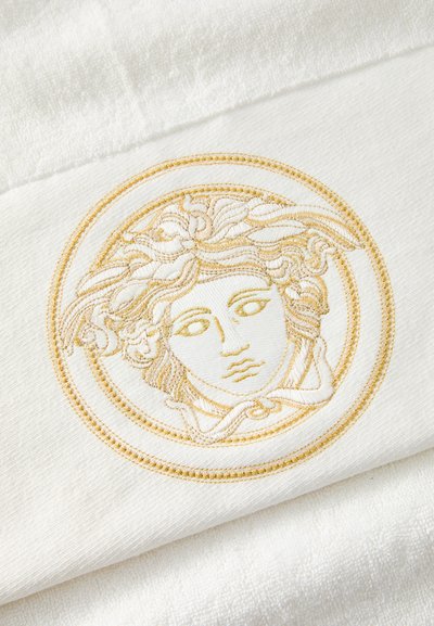 Prosop alb cu o broderie aurie proeminentă reprezentând un chip în interiorul unei margini circulare, evidențiind detalii complicate și țesătură texturată.