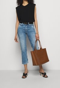 Braune Ledertasche mit geprägtem Logo, schwarzes ärmelloses Oberteil, hellblaue cropped Jeans und schwarze High Heels mit Knotendetail.