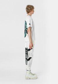 Camiseta blanca con un gran diseño de alas de mariposa turquesa y negra en la parte posterior, combinada con pantalones blancos que presentan acentos gráficos en blanco y negro.