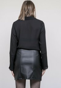 Chemise noire à manches longues avec des poignets plissés, associée à une mini-jupe en cuir noir taille haute, vue de dos.