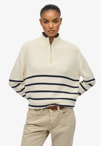Jersey cremoso con cremallera y rayas marinas, cuello acanalado y textura suave. Combinado con pantalones beige claros y un cinturón trenzado marrón.