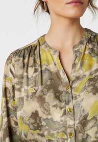 Frau mit einer Bluse in grünem, beigem und gelbem abstraktem Muster, die einen Mandarin-Kragen und gelbe Knöpfe hat. Abgebildet von den Schultern bis zum Kinn.