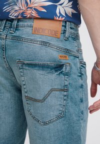 Un jean en denim bleu clair arbore une patte en cuir marron portant l'inscription "INDICODE", une poche arrière sculptée et des surpiqûres contrastantes le long des coutures.