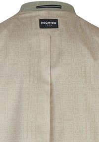 Beige jacket met een subtiele all-over 'Hechter' logo patroon. Opvallende puntkraag met contrasterende stof en een achterste middelnaad.