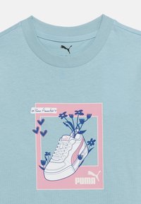 Lyseblå Puma T-shirt med en grafik af en hvid sneakers fyldt med blå blomster inde i en lyserød firkantet ramme.