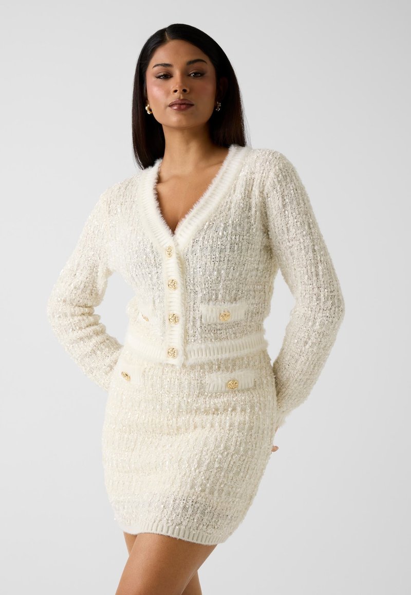 Robe cardigan en maille blanche duveteuse avec un col en V profond, des boutons dorés et des poches avant ; texture côtelée et légère brillance.