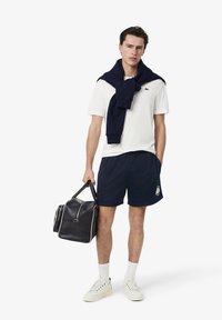 Shorts atléticos navy, camiseta blanca con logo, suéter navy colgado, llevando un bolso de lona texturizado navy y usando zapatillas blancas con detalles.