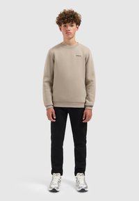 Tienerjongen staat rechtop in een beige sweatshirt, zwarte broek en witte sneakers tegen een simpele grijze achtergrond.