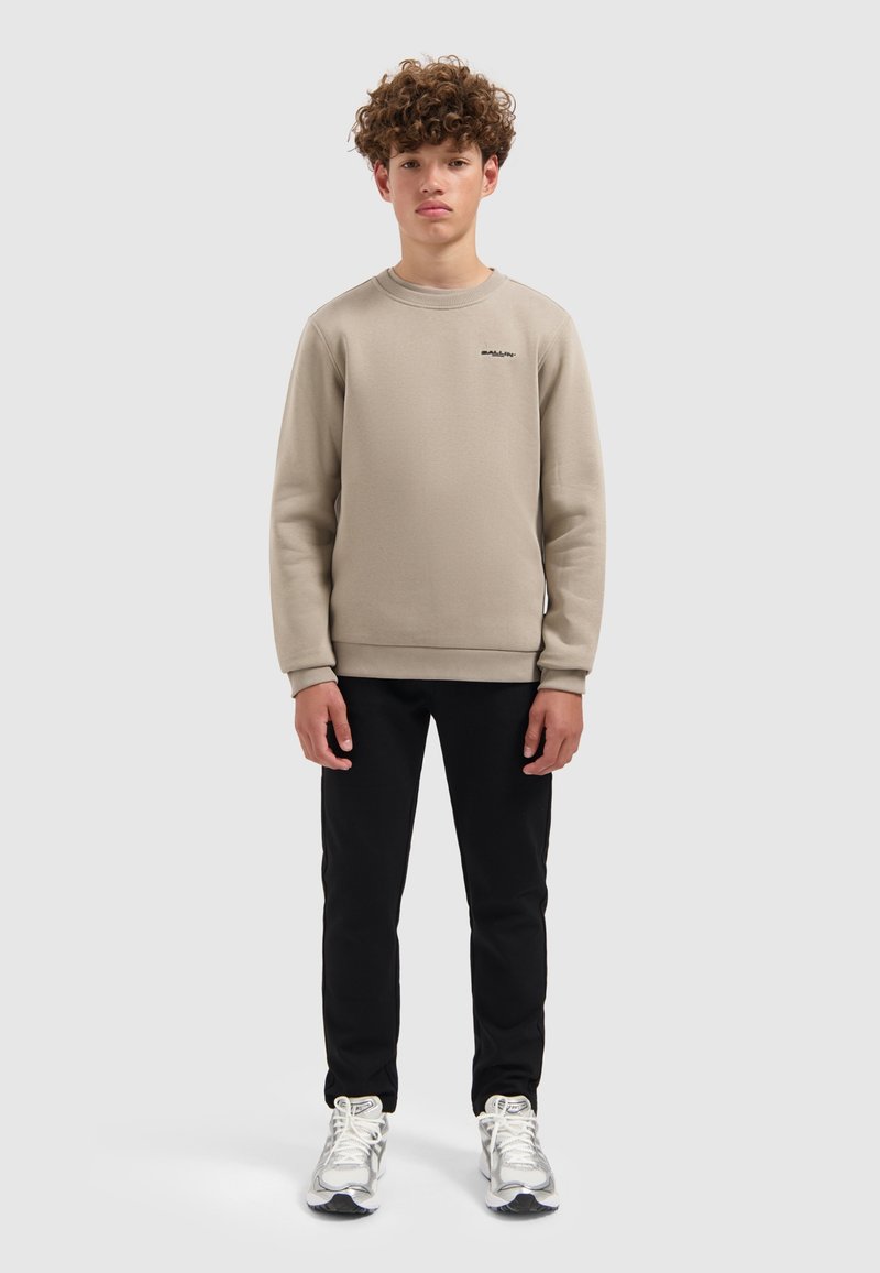 Tienerjongen staat rechtop in een beige sweatshirt, zwarte broek en witte sneakers tegen een simpele grijze achtergrond.