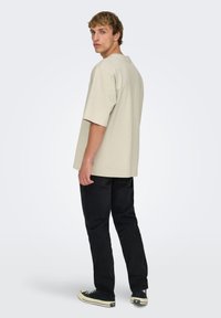 T-shirt beige à manches courtes avec une coupe décontractée, un col rond et des fentes latérales, associé à un jean noir et des baskets noires.