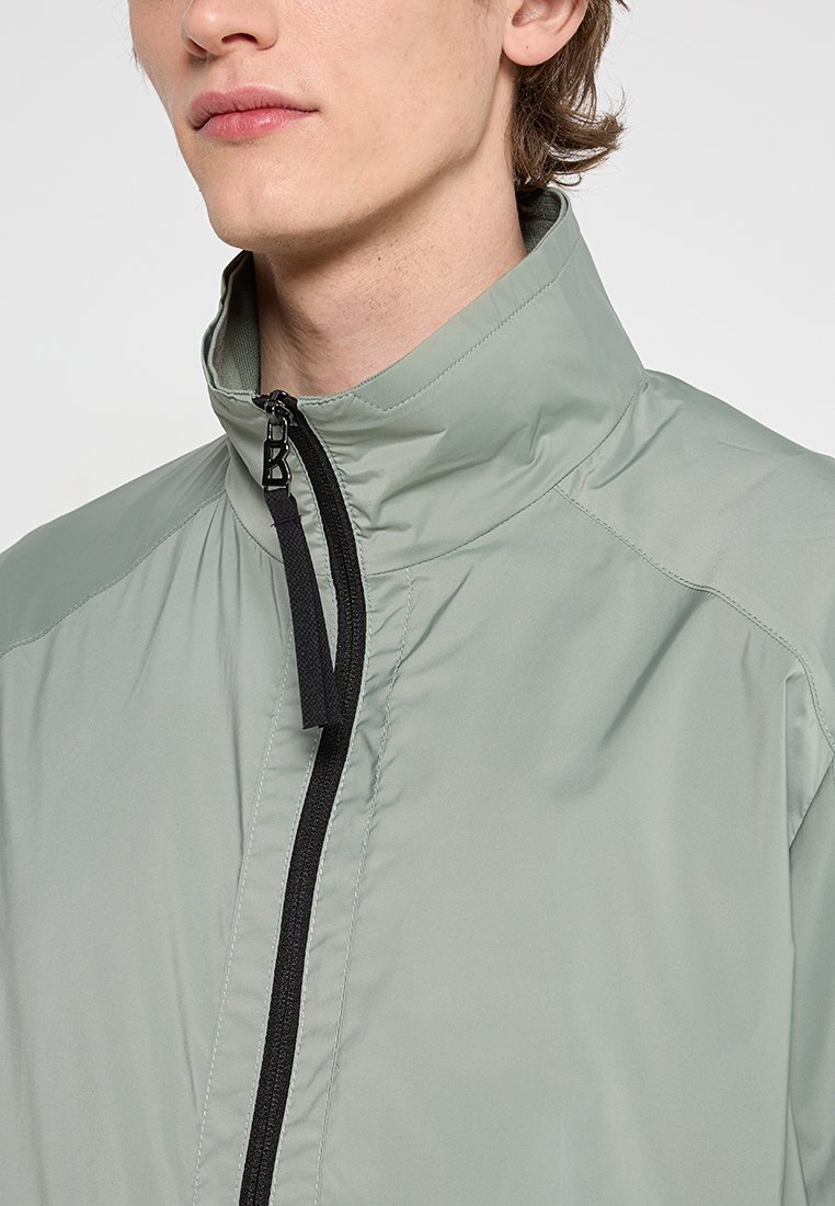 Hellgrüne, leichte Jacke mit hohem Kragen, durchgehendem Reißverschluss und schwarzen Reißverschlussdetails. Glatte Textur und minimalistisches Design.