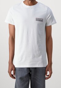 Bílý bavlněný tričko s kulatým výstřihem, krátkými rukávy a obdélníkovou nášivkou na horní levé straně hrudi. Pohodlný střih, minimalistický design.