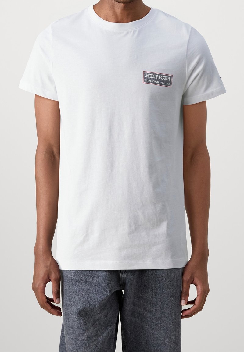 Bílý bavlněný tričko s kulatým výstřihem, krátkými rukávy a obdélníkovou nášivkou na horní levé straně hrudi. Pohodlný střih, minimalistický design.