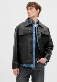 Sort læderjakke med store brystlommer, knaplukning og spidse krave, lagdelt over en denimskjorte. Grå jeans fuldender looket.
