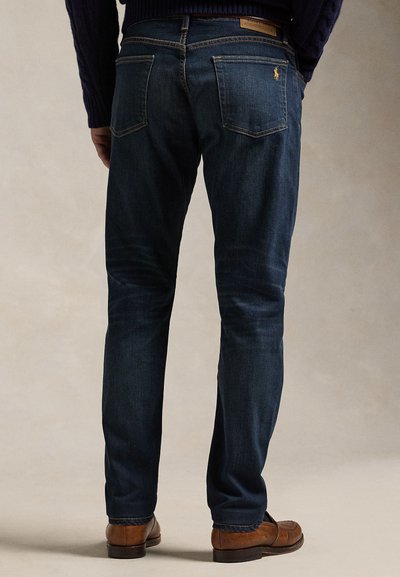 Muškarac odjeven u tamnoplave uske traperice, smeđe kožne cipele i tamni džemper, prikazan s leđa na neutralnoj pozadini.