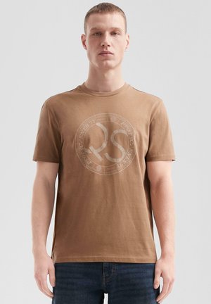 Bruine katoenen t-shirt met een ronde hals. Heeft een groot, lichtgekleurde grafisch logo in het midden. Casual pasvorm, korte mouwen.