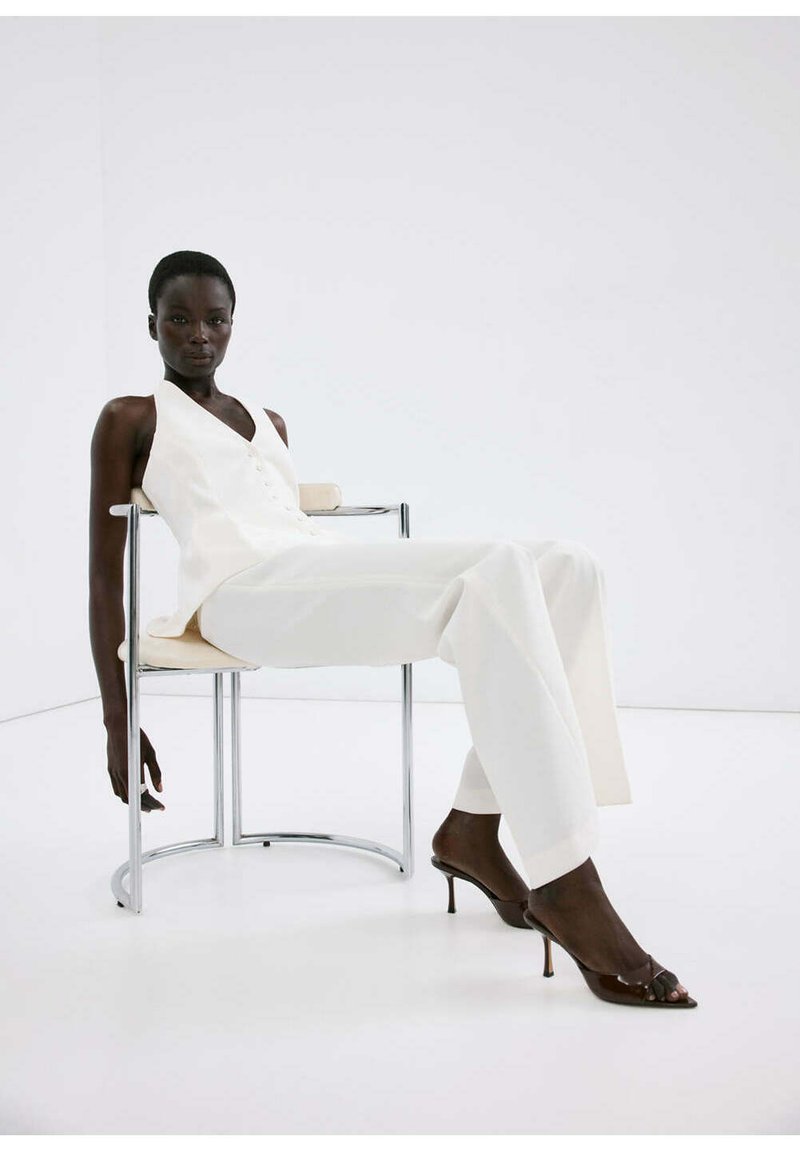 Femme en haut blanc sans manches et pantalon, assise détendue sur une chaise métallique moderne, portant des talons ouverts foncés dans une pièce lumineuse et minimaliste.