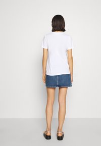 Bílé bavlněné tričko s krátkými rukávy, zobrazené zezadu. Kombinováno s džínovou sukní s neupraveným lemem. Viditelné černé sandály.