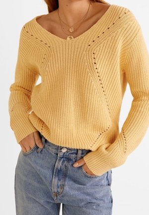 Woman wearing a yellow knit V-neck sweater and light blue denim jeans with hands in pockets, accessorized with layered gold necklaces.