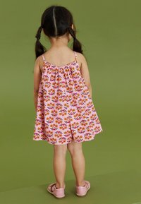 Combi-short rose à imprimé floral avec des fines bretelles, un col froncé et une coupe évasée et décontractée. Associé à des sandales roses, présenté sur un fond vert.