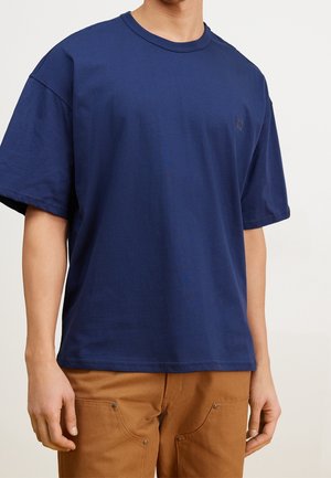 T-shirt en coton bleu marine à manches courtes, col rond et petit patch logo discret sur la poitrine. Texture douce, coupe décontractée.