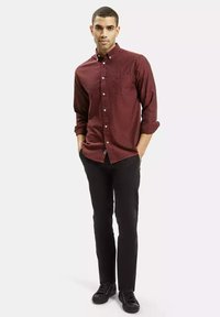 Bordowa koszula z długim rękawem i zapięciem na guziki, z kieszonką, w połączeniu z czarnymi spodniami i czarnymi butami. Koszula ma gładką fakturę oraz podwinięte rękawy.