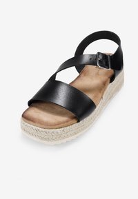Sandale compensée en cuir noir avec des brides croisées, une semelle en suède beige et une semelle en jute. Détail de boucle en métal réglable.