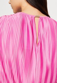 Różowa plisowana bluzka z wycięciem na plecach oraz metalicznym akcentem przy dekolcie, prezentująca gładki, zwiewny materiał w intensywnym kolorze.