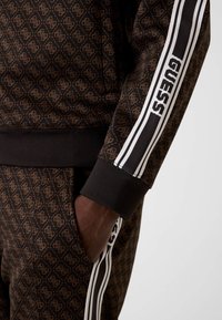 Brązowy dres z wzorzystym, fakturowanym designem. Z białymi paskami na rękawach i ściągaczami przy mankietach. Po bokach widoczna kieszeń.