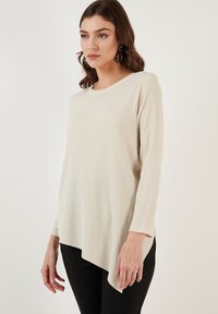 Top beige a maniche lunghe in tessuto testurizzato, scollo a girocollo e orlo asimmetrico. Abbinato a pantaloni neri.