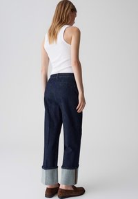 Jean en denim bleu foncé taille haute avec des ourlets retroussés révélant une bande de denim plus clair, associés à un top sans manches blanc et des chaussures plates marron.