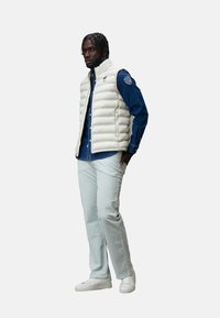 Gilet imbottito in bianco sporco, con design a righe orizzontali, abbinato a una camicia blu scuro, pantaloni verde chiaro e sneakers bianche.