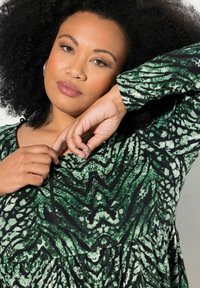 Vihreä kuvioitu mekko, jossa on tekstuuriin perustuva käärmeenkaltainen muotoilu. Mekossa on muotoiltu yläosa ja hulmuava alaosa sekä musta vetoketjulisäys kaula-aukossa.