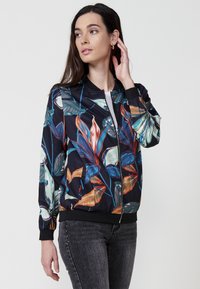 Giacca bomber floreale in tessuto scuro, raffreddato da stampe colorate di foglie. Chiusura con zip, polsini e orlo a costine, e tasche su entrambi i lati.