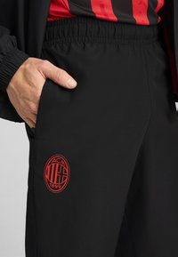 Pantalons de sport noirs avec taille élastique, arborant un logo rouge ACM 1899 et des poches latérales. Texture lisse, design à jambe droite.