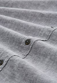 Chemise en lin grise avec plusieurs plis, dotée de boutons noirs sur le devant. La texture et les détails de couture sont visibles.