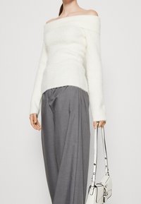 Maglione bianco a spalle scoperte, morbido e lavorato a maglia, con maniche lunghe, abbinato a pantaloni grigi larghi e con in mano una piccola borsa bianca con dettagli neri.