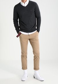 Pull gris foncé à col en V porté sur une chemise blanche à col, associé à un pantalon chino beige et des baskets blanches. Détails remarquables : poignets retroussés et marquage de la marque.