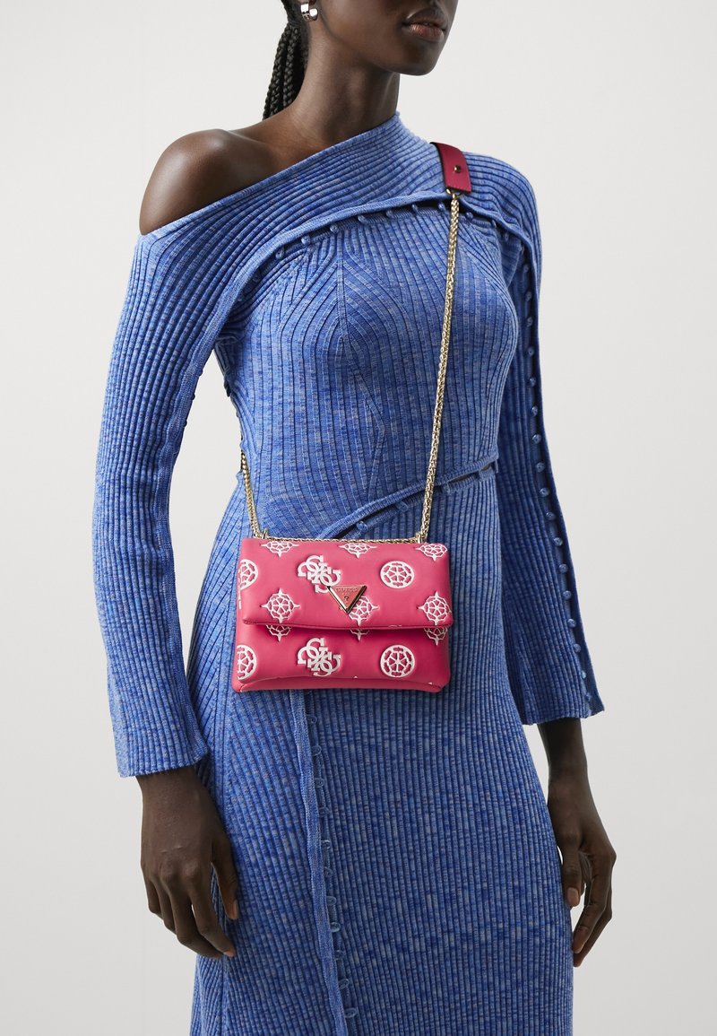 Femme portant une robe bleue côtelée épaules dénudées avec un sac bandoulière rose à motif avec une lanière chaîne dorée.