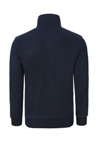 Tmavě modrá fleecová bunda s vysokým límcem, žebrovanými manžetami a spodním lemem. Na zádech je vyražený text "POLO CLUB LA MARTINA 1987".