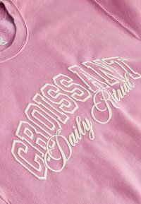 Light pink sweatshirt featuring white graphic text reading "CROISSANT Daily Ritual" with bold and cursive font styles. Soft fabric texture.