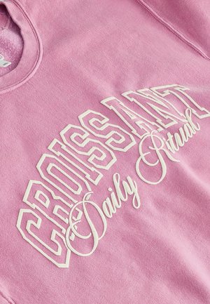 Light pink sweatshirt featuring white graphic text reading "CROISSANT Daily Ritual" with bold and cursive font styles. Soft fabric texture.