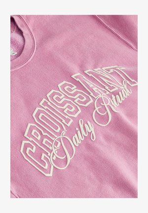 Light pink sweatshirt featuring white graphic text reading "CROISSANT Daily Ritual" with bold and cursive font styles. Soft fabric texture.