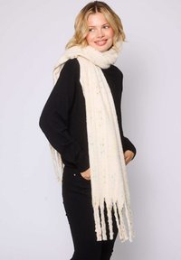 Femme blonde portant une tenue noire et une longue écharpe crème avec des specks multicolores et des franges, souriante et regardant sur le côté.