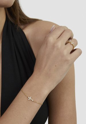 Woman wearing a black top with a gold bracelet and gold ring, both featuring cross-shaped diamond designs.