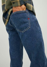 Jeans de denim azul con un corte recto, costuras visibles y una etiqueta roja en el bolsillo trasero. La textura es suave con un ligero brillo.