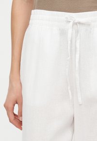 Pantaloni albi din in cu talie elastică și șnur legat, purtați cu un top bej, mâna odihnindu-se lângă pantaloni.