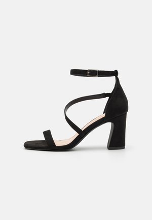 Black suede heeled sandal featuring a square toe, ankle strap with a metal buckle, and crossed straps over the foot. Block heel design.