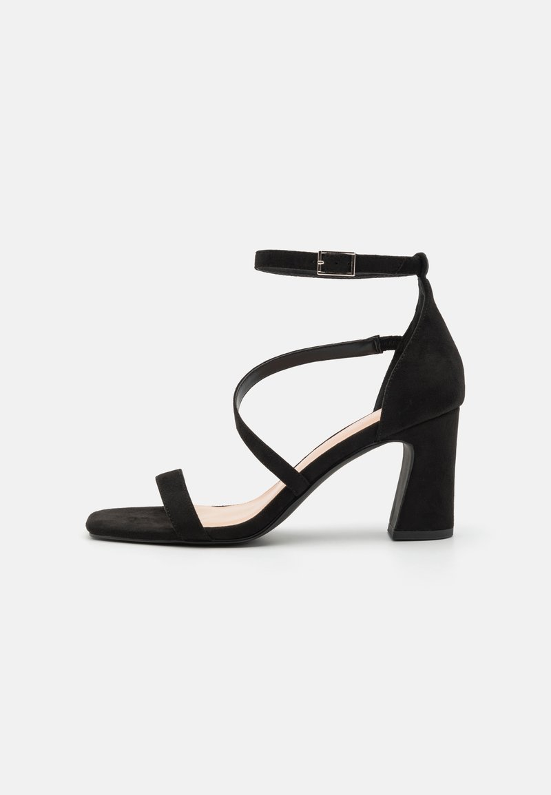Sandale à talon en daim noir avec bout carré, bride de cheville avec boucle en métal et brides croisées sur le pied. Design à talon bloc.